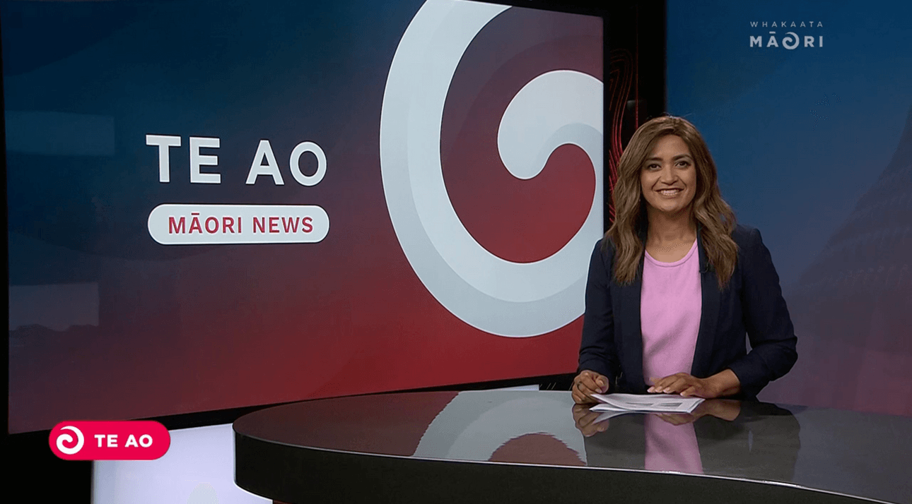 A news anchor sitting at a desk in a studio with a "Te Ao Māori News" backdrop. The person is wearing a pink top and dark blazer, smiling at the camera. The studio has a dark red and blue colour scheme with a large spiral design.